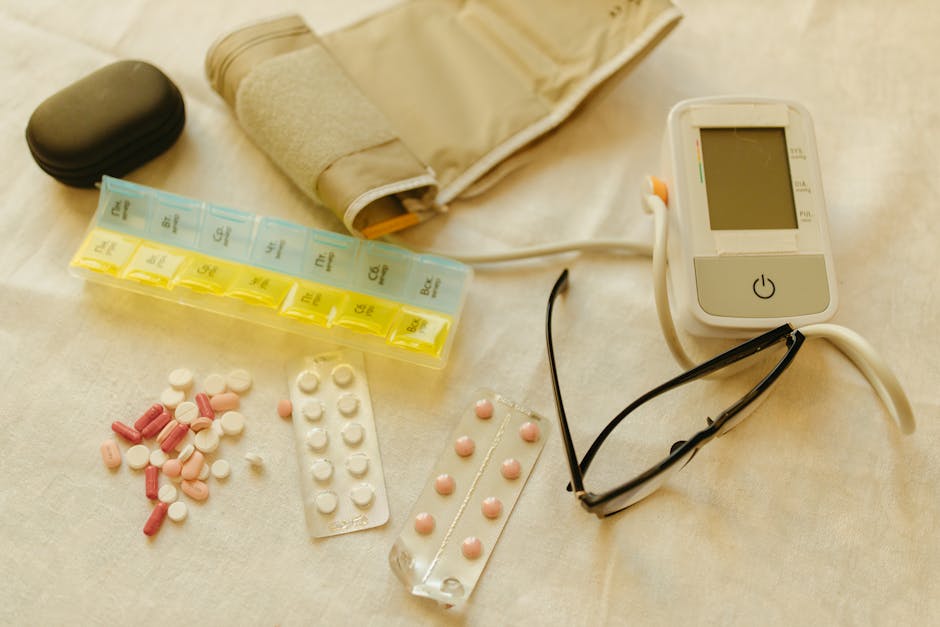 Flat lay image of healthcare essentials including a blood pressure monitor, pills, and eyeglasses on a white surface.